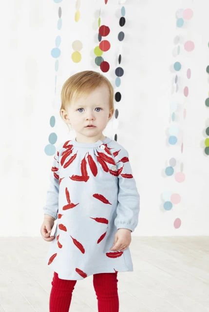 Toddler girl in light blue dress with feather print and red leggings standing indoors against white wall with colorful polka dot garlands.