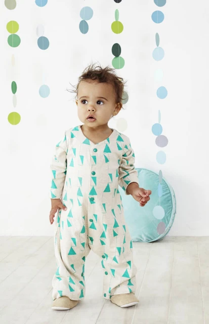 A baby stands indoors in a patterned jumpsuit, looking to the left, with blue and green decorations hanging in the background.