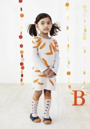 Full-length portrait of a young girl with pigtails in a feather-patterned dress and knee-high socks, standing in a brightly lit interior.