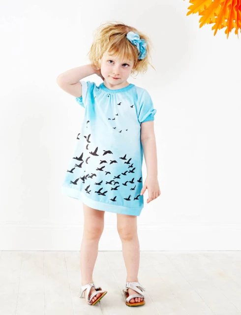 Adorable young girl with blonde hair poses in a light blue dress with birds printed on it in a bright, white studio setting. 