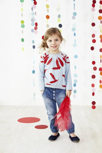 A young girl with blonde hair, wearing a light blue long-sleeved shirt and jeans, smiles while holding a red feather in a studio setting.