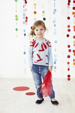 A young girl with blonde hair, wearing a light blue long-sleeved shirt and jeans, smiles while holding a red feather in a studio setting.