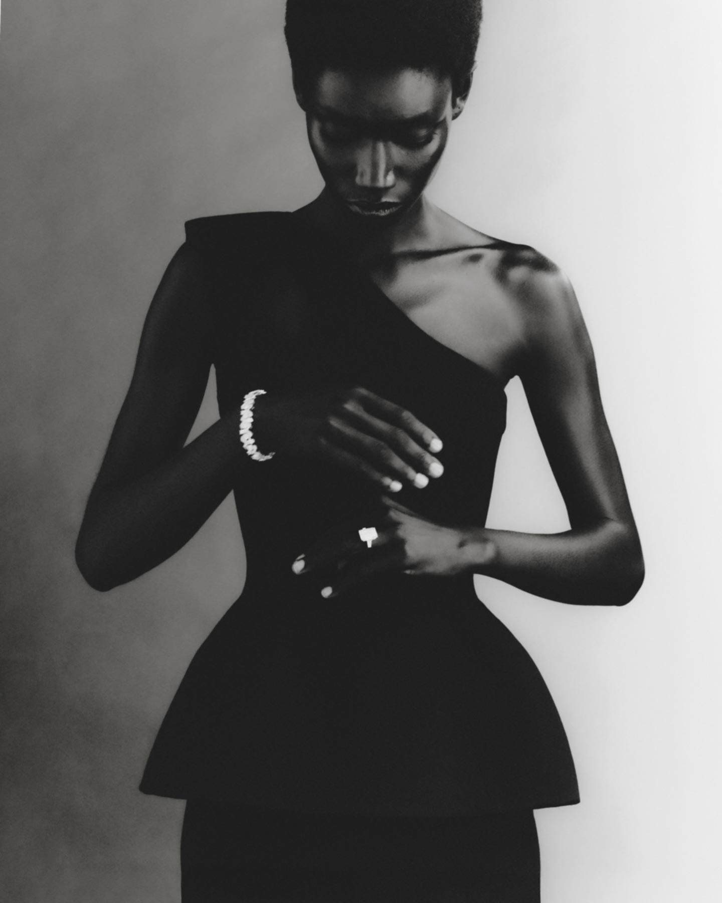 A slender Black woman wearing a one-shoulder black dress and jewelry in a studio setting, looking down with a neutral expression.