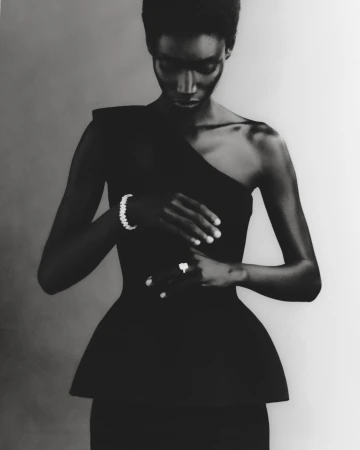 A slender Black woman wearing a one-shoulder black dress and jewelry in a studio setting, looking down with a neutral expression.