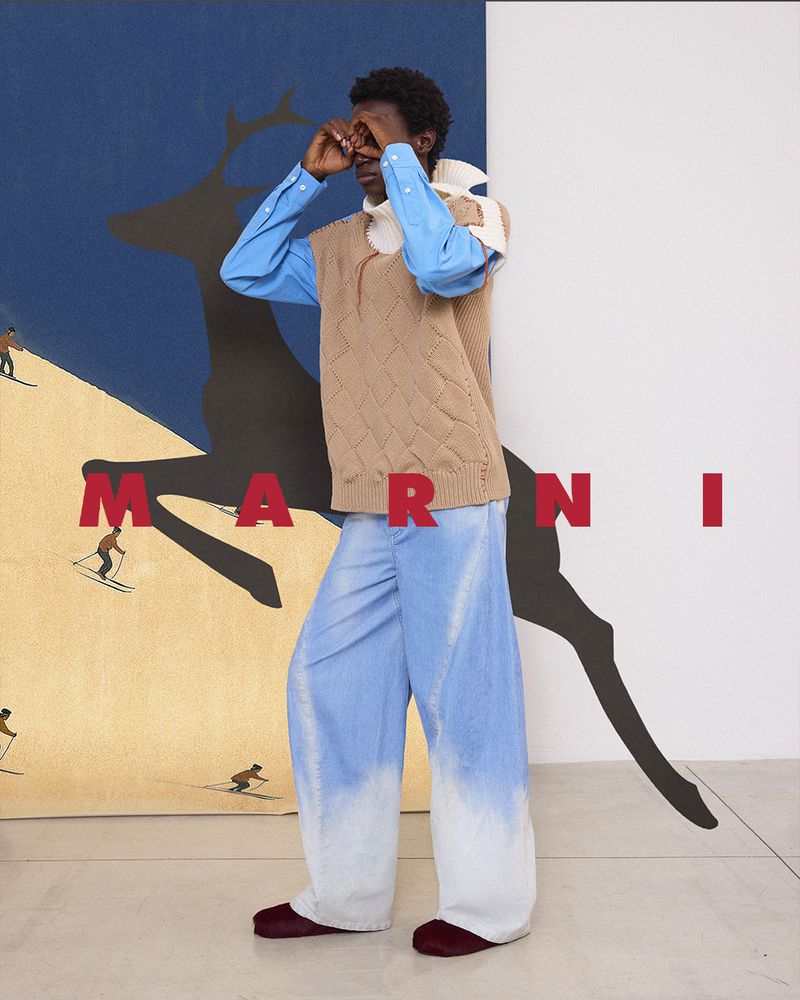 A dark-skinned man in a sweater vest poses with hands framing his eyes, standing in front of a painting for a fashion editorial.