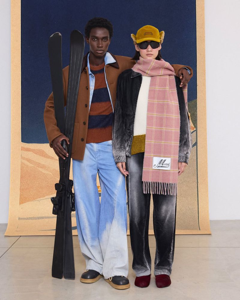 Two fashion models pose indoors against a backdrop, one holding skis, both wearing coats, sweaters, and trousers in a studio setting.