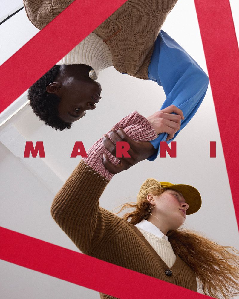 Fashion editorial shot from a low angle featuring two models wearing knitwear and set against a red and white background with text.