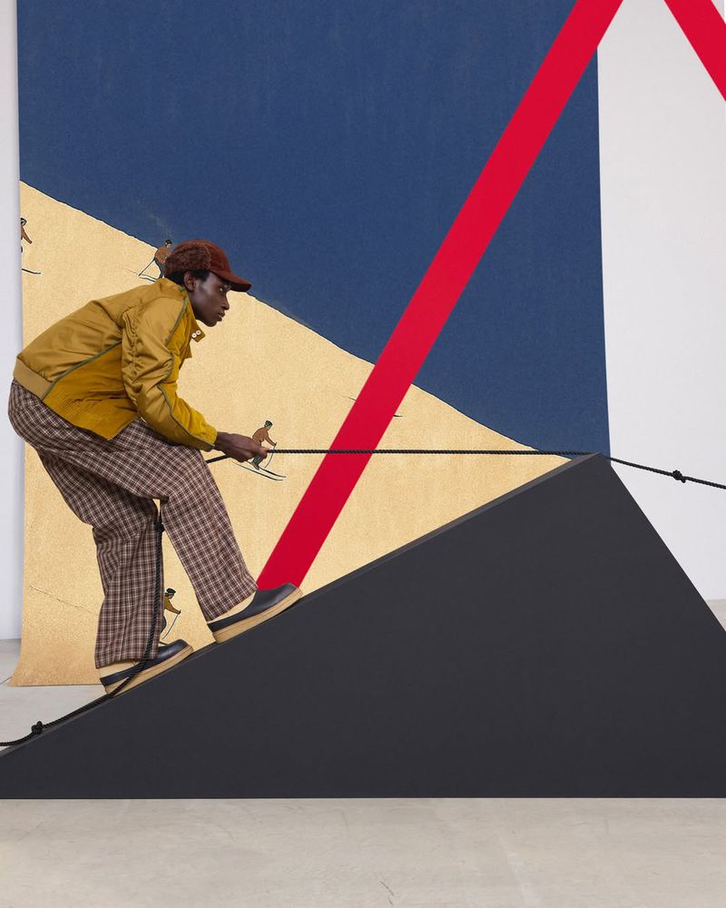 Person in a brown cap and yellow jacket appears to be skiing on a black wedge, with a stylized backdrop and miniature skiers.