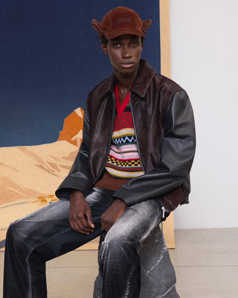 Studio shot of a Black male model wearing a brown cap, jacket, patterned sweater, and distressed jeans against a landscape painting.