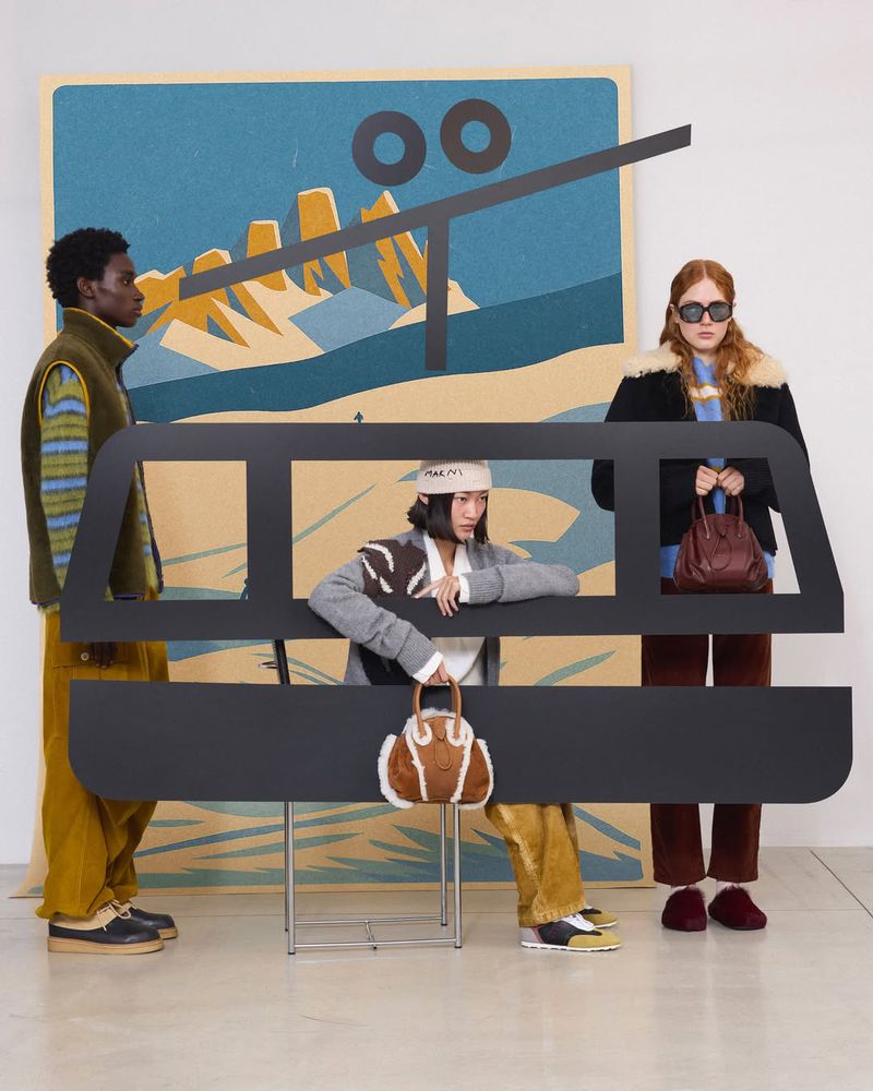 Three models pose behind a prop of a ski lift in a studio. The background features a mountain range and a blue sky illustration.