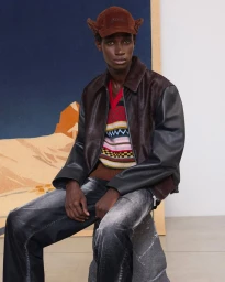 Model wearing brown cap, patterned knit vest, dark jacket and jeans seated in front of a painted backdrop for Marni.