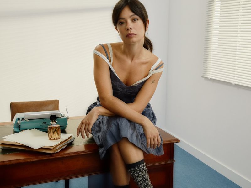 Brunette woman in sundress poses on a desk near a typewriter. She is looking at the camera with a neutral facial expression.