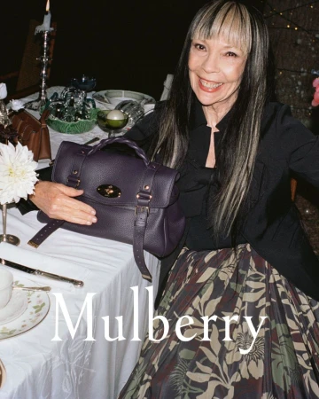 Elderly woman smiles with a purple Mulberry handbag on a table set with dinnerware and silverware in a dark indoor setting.