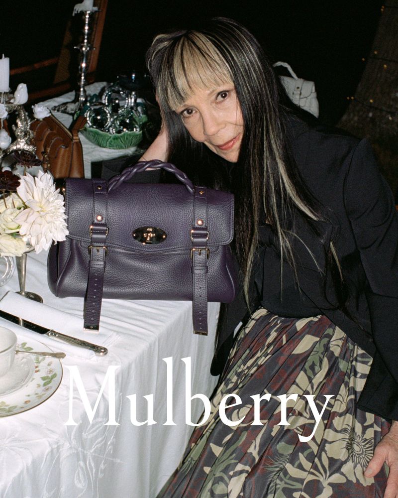 Brunette woman poses with a purple leather purse in front of a set table with floral arrangements and white tablecloths.