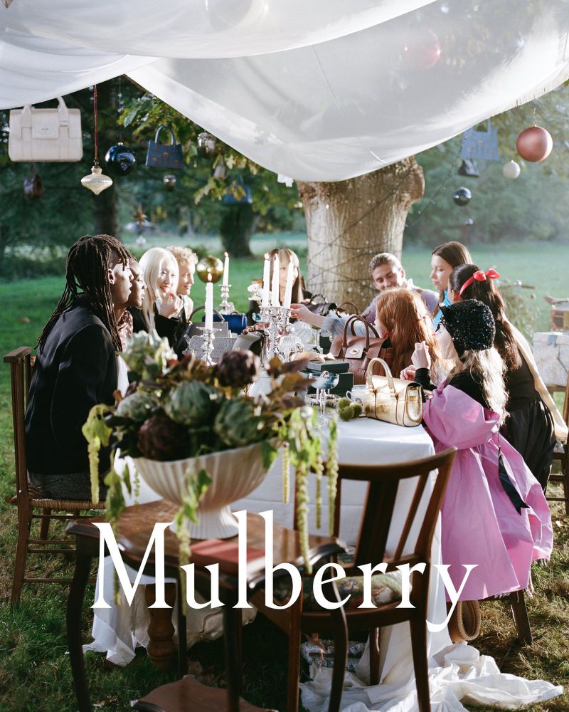Outdoor fashion editorial shows a group of adults and children sitting at a table decorated with flowers and hanging handbags.