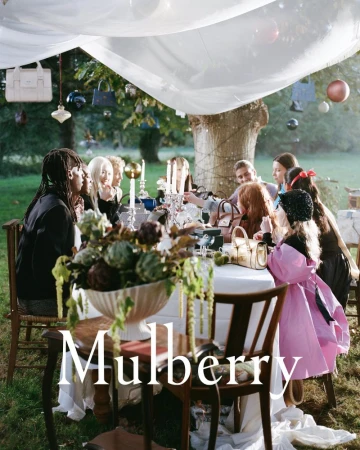 Outdoor fashion editorial shows a group of adults and children sitting at a table decorated with flowers and hanging handbags.