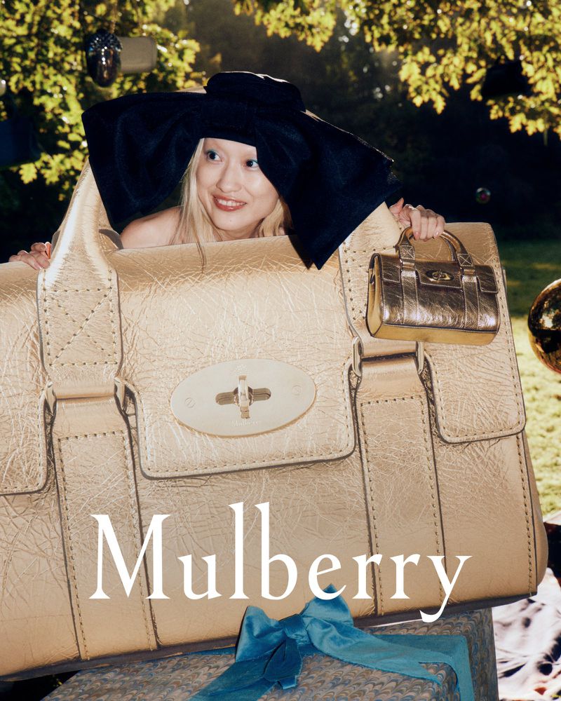 Fashion shot featuring a fair-skinned adult female wearing a black hat holding two metallic gold bags, displayed outdoors in natural light.