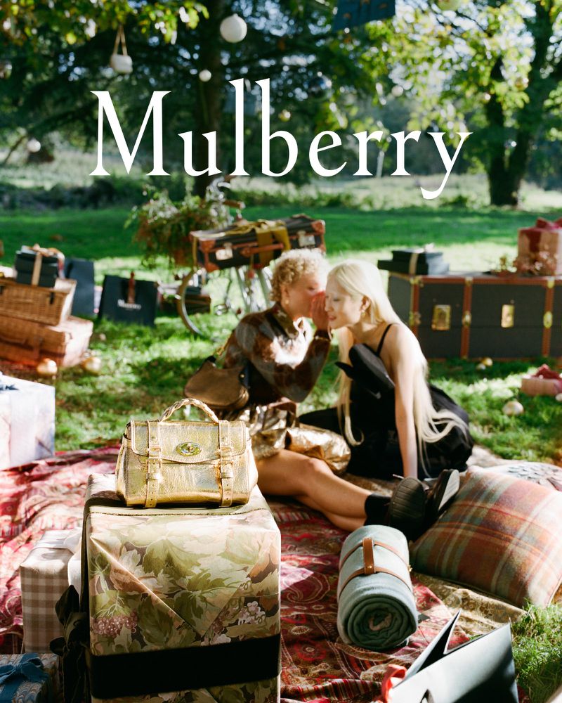 Two women are picnicking in a grassy, sunlit clearing, surrounded by vintage luggage and presents, promoting the 'Mulberry' brand.
