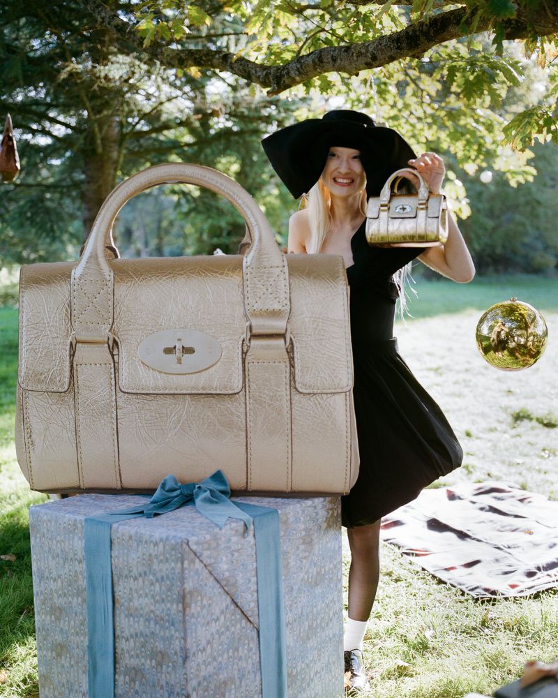 Large gold-colored purse resting on a gift box outdoors, woman in hat holds a smaller matching purse, trees in background.
