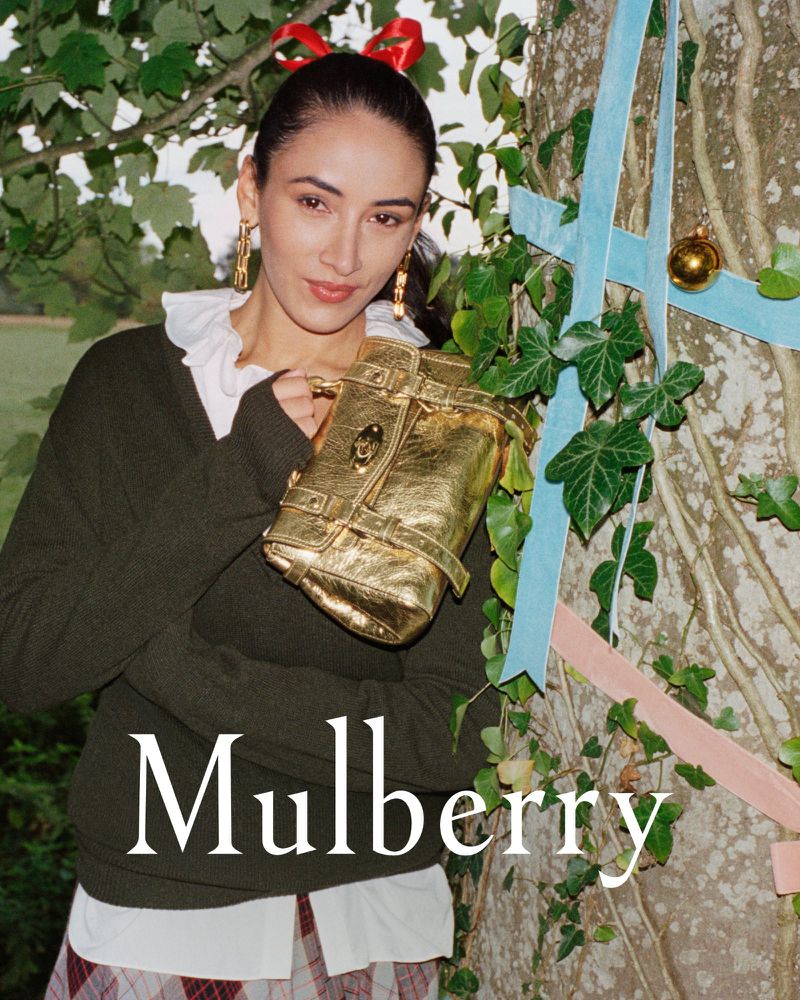 A woman poses with a gold Mulberry handbag outdoors, wearing a green sweater, and bow in her hair with a decorative tree in the background.