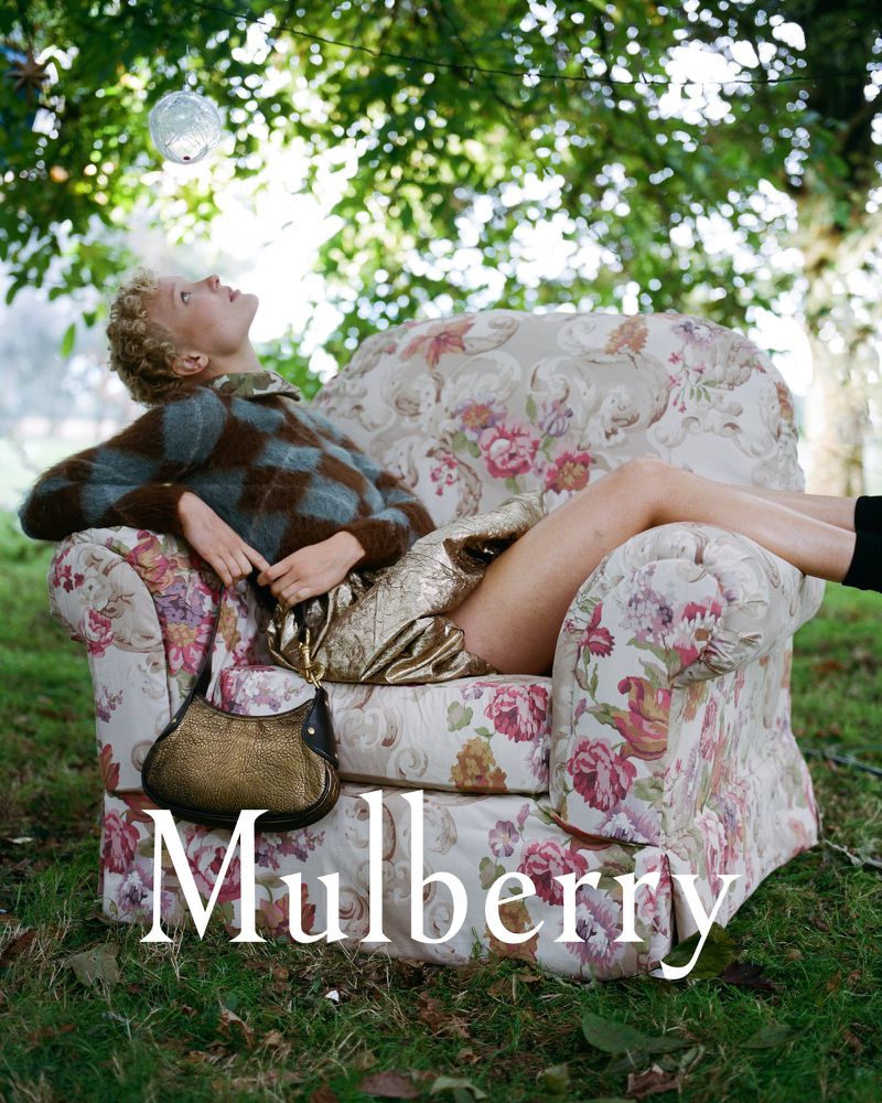 A woman in a patterned sweater rests on a floral armchair outside, holding a gold handbag, under a tree with a mirrored ball hanging.