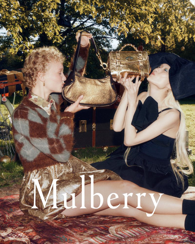 Two adult women model golden handbags for a fashion campaign in a grassy outdoor setting with trees, a patterned blanket, and text overlay.