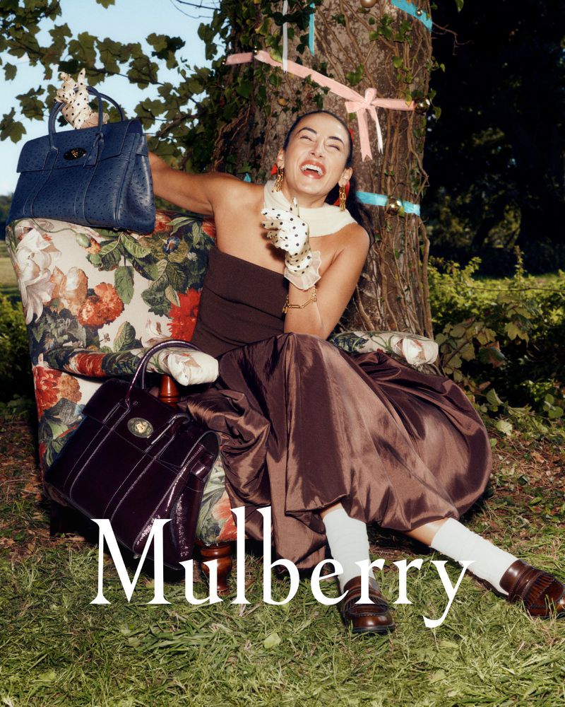Fashion shot of a woman in a brown dress and white socks, seated on a floral chair with Mulberry handbags in a grassy outdoor setting.