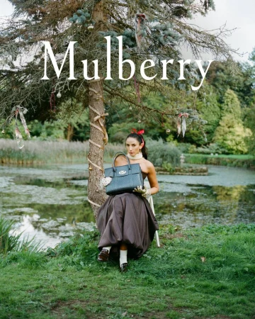 Model seated under an outdoor decorated tree by a pond holding a dark Mulberry leather handbag in a countryside garden setting.