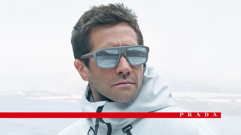 A man poses wearing Prada sunglasses and a light-colored hooded jacket with a bright white and grey background. The Prada logo is visible.