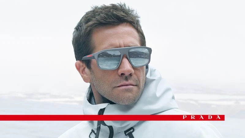 A man poses wearing Prada sunglasses and a light-colored hooded jacket with a bright white and grey background. The Prada logo is visible.