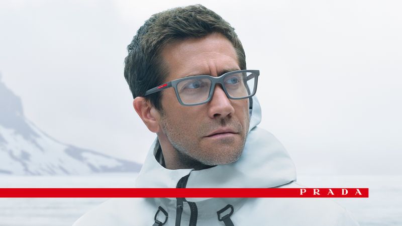 Man wearing gray Prada glasses in an outdoor setting with snow-covered mountains; red Prada logo text at the bottom of the frame.