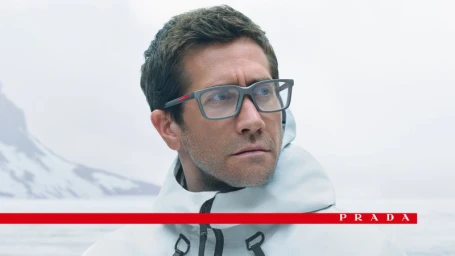 Jake Gyllenhaal wearing grey rectangular eyeglasses and a white hooded technical jacket against a snowy glacier background.