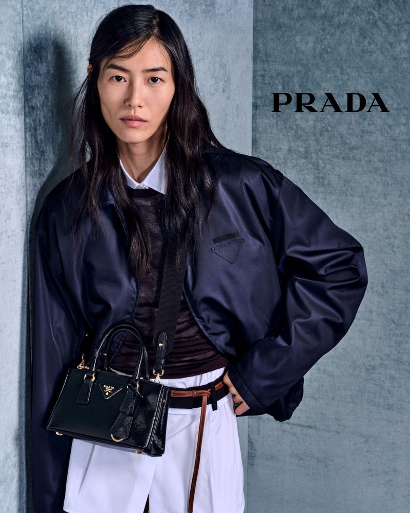 Fashion model wearing Prada navy bomber jacket with black leather handbag against grey wall with Prada logo in studio shot.