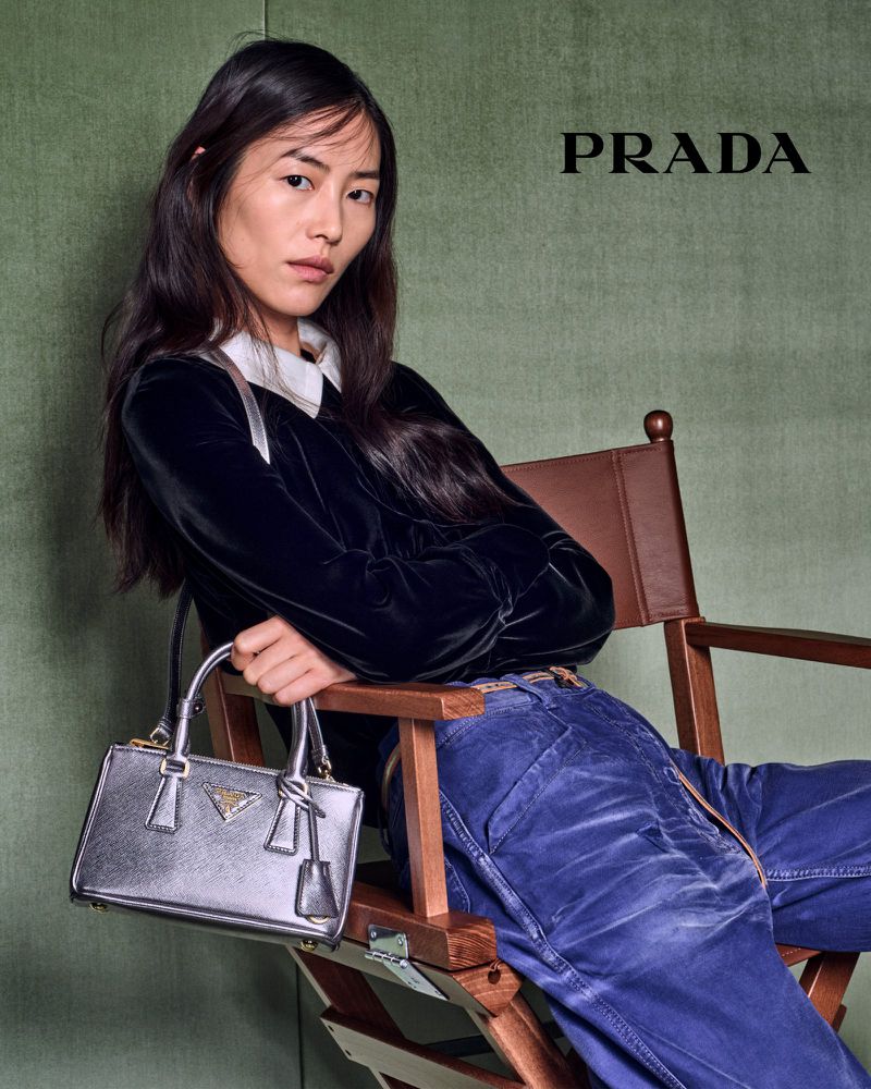 A woman poses indoors in velvet top and blue jeans, seated in a wood chair with a silver Prada handbag. Fashion photography.