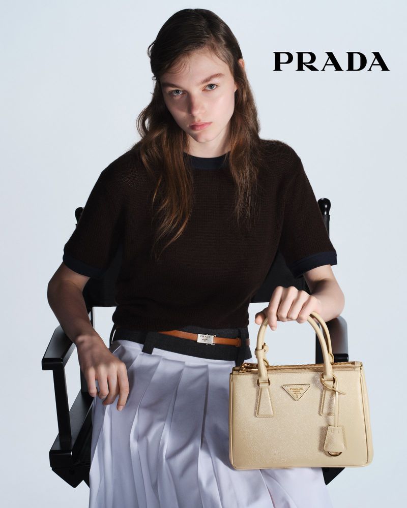 Fashion shot featuring a fair-skinned model sitting in a director's chair, holding a tan Prada purse in front of a white backdrop.