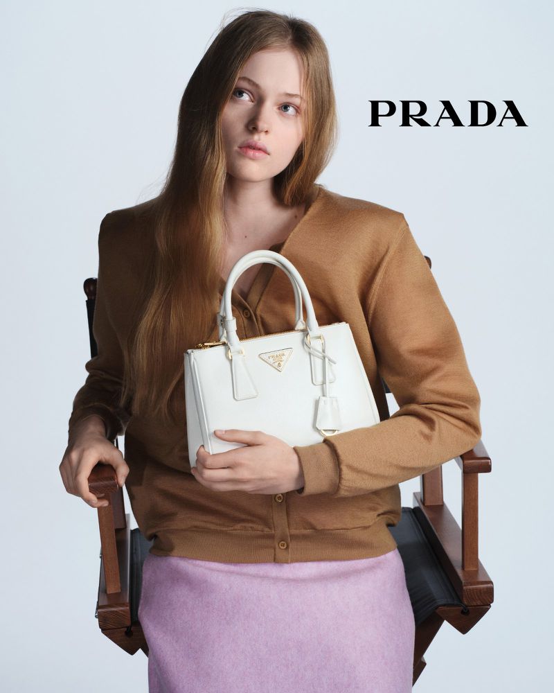 Indoor shot of a fair-skinned young woman in a tan cardigan holding a white Prada purse while sitting on a wooden chair.