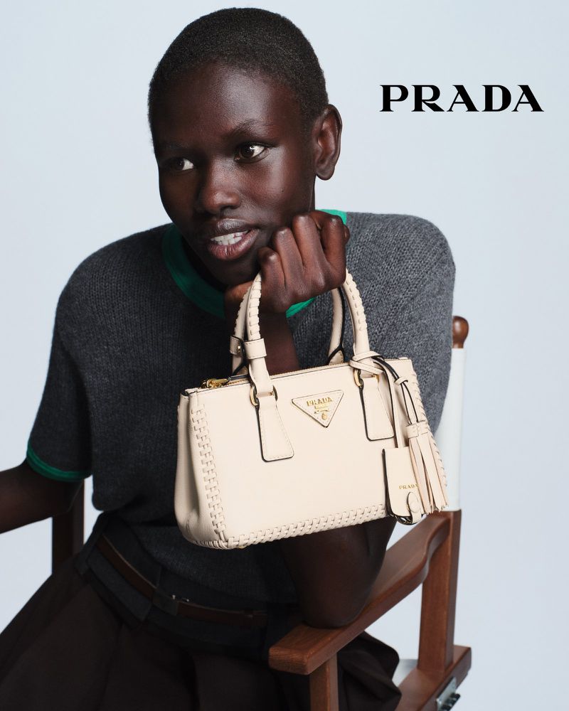 Close-up shot of an African woman sitting with a cream-colored Prada handbag in neutral studio lighting. Fashion and beauty advertising.