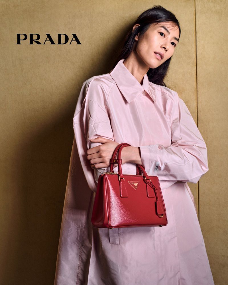 Asian woman in a pink coat holds a shiny red Prada handbag against a textured gold background in a studio product shot.