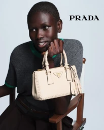 Rejoice Chuol seated on a director's chair holding a cream Prada handbag against a pale studio backdrop.