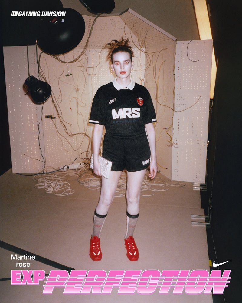 A woman poses indoors wearing a soccer jersey and shorts with red sneakers, against a backdrop of studio lighting and wires.