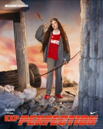 Model wearing a grey tracksuit, red MRS jersey, and crown while holding a bow and arrow amidst classical stone ruins and rubble.