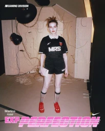 Model wearing a black MRS football jersey, matching shorts, and red athletic shoes on a set with exposed wires.