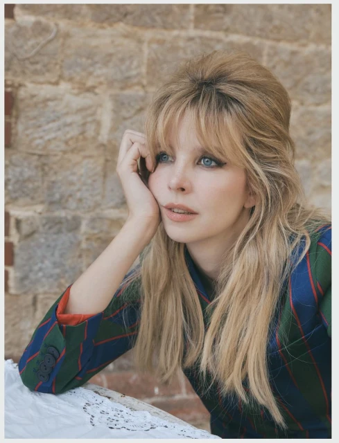 Portrait of a blonde woman with blue eyes in a plaid blazer, resting her head on her hand in front of a stone brick wall.