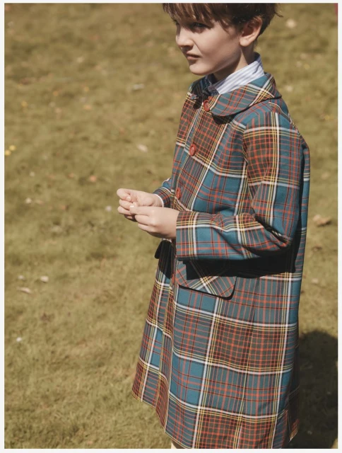 A young boy stands outdoors wearing a long plaid coat, with a blue striped collared shirt and a bow tie, with a field in the background.