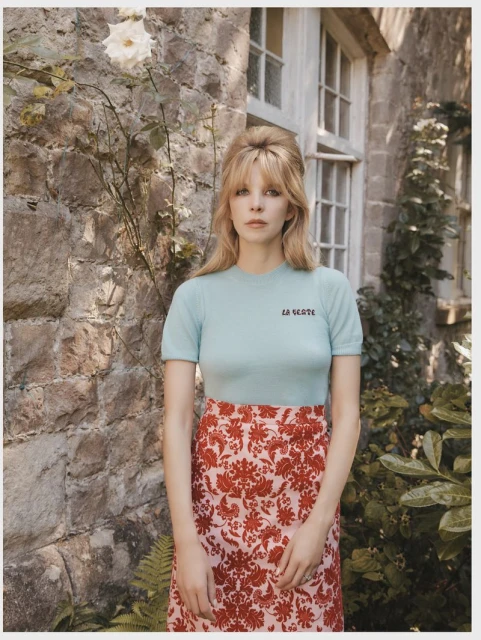 Blonde woman in a light blue top and floral skirt stands in front of a stone wall and window with a rose bush next to the wall.