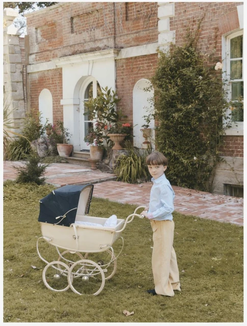 A young boy stands on a lawn in front of a brick building pushing a vintage-style cream-colored pram with a dark blue hood.