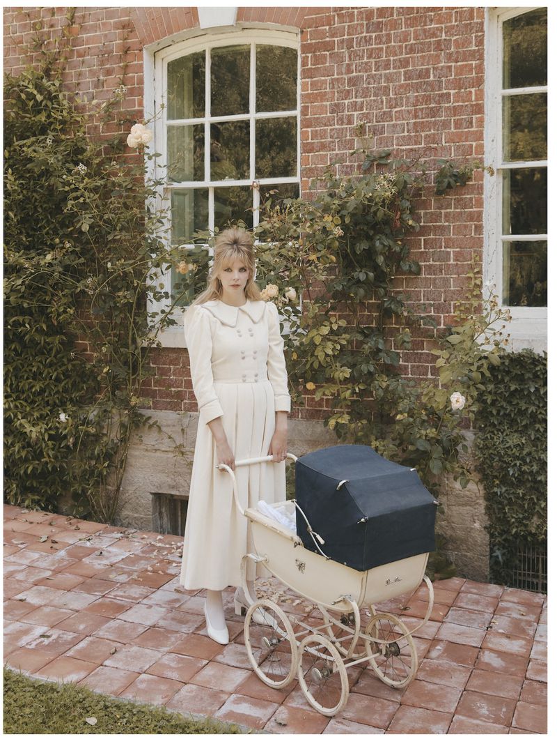 An adult female poses outdoors in a long, light-colored dress, standing next to a vintage baby stroller on a brick-paved area.