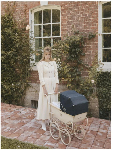 An adult female poses outdoors in a long, light-colored dress, standing next to a vintage baby stroller on a brick-paved area.