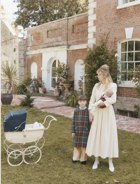 A woman holding a baby stands next to a boy and a baby carriage outside a brick building on a lawn during the daytime.