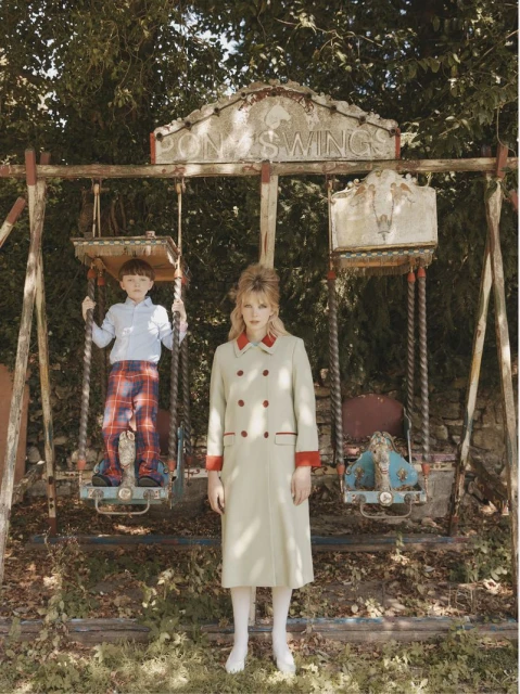 An adult female in a beige coat and a child in a blue shirt and plaid pants pose next to a vintage swing set surrounded by lush trees.